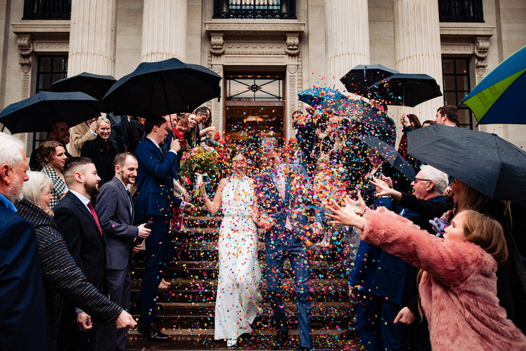 A couple walking through a field, with confetti thrown by their guests at Old Marylebone Town Hall.