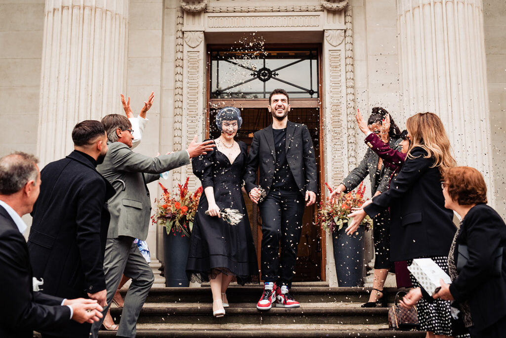 A bride and groom descend the steps of Marylebone Town Hall, while their wedding guests throw confetti around them. 