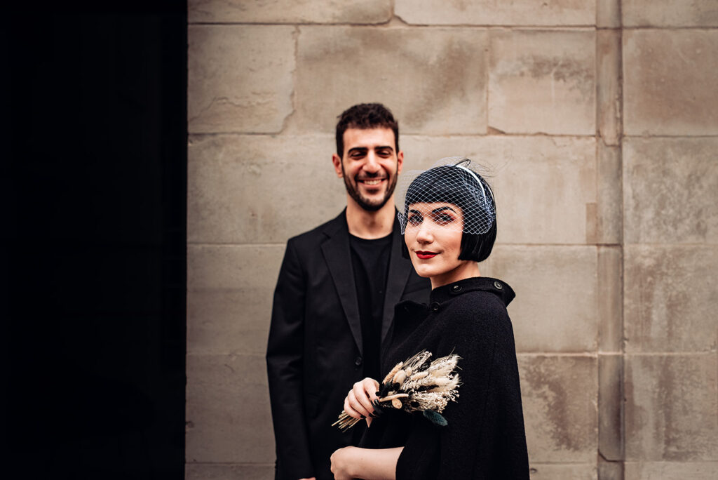 A bride and groom, on their wedding day, dressed in black. She holds a bouquet of dried flowers. 