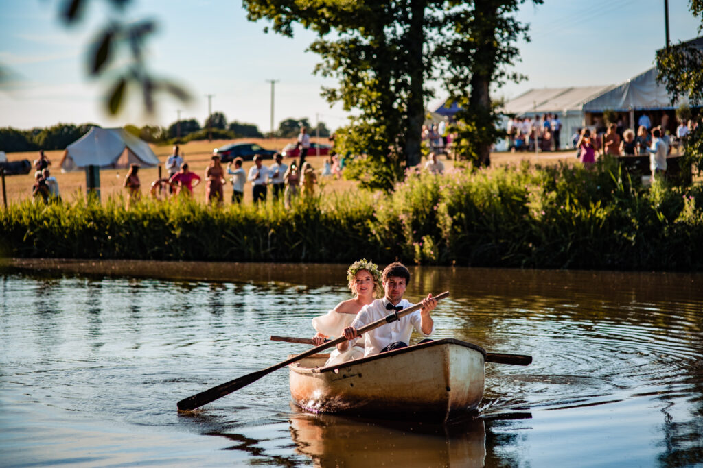A bride and groom on their wedding day, taking time to row a small boat on a river. Behind them out of focus is an outdoor wedding with tents, with all the guests scattered outside it in the sun.