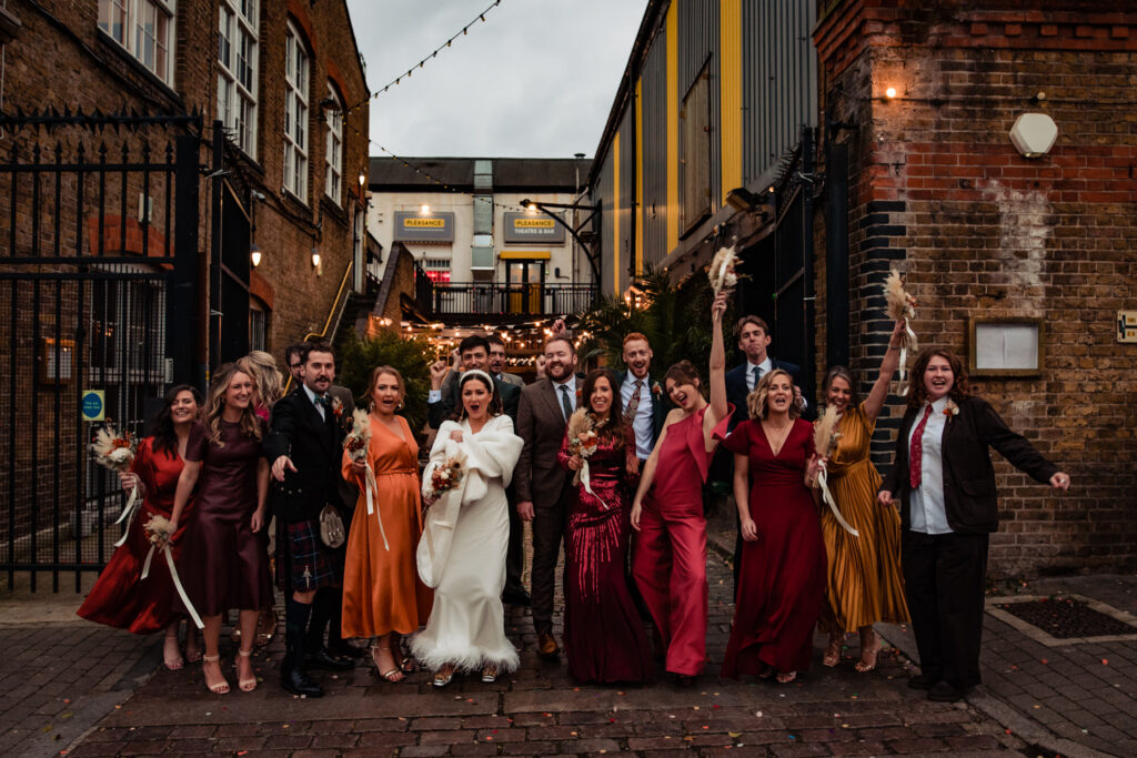 A 2022 wedding party cheering in an outdoor, industrial setting. The colours are warm autumnal and wintery reds, oranges, and browns.