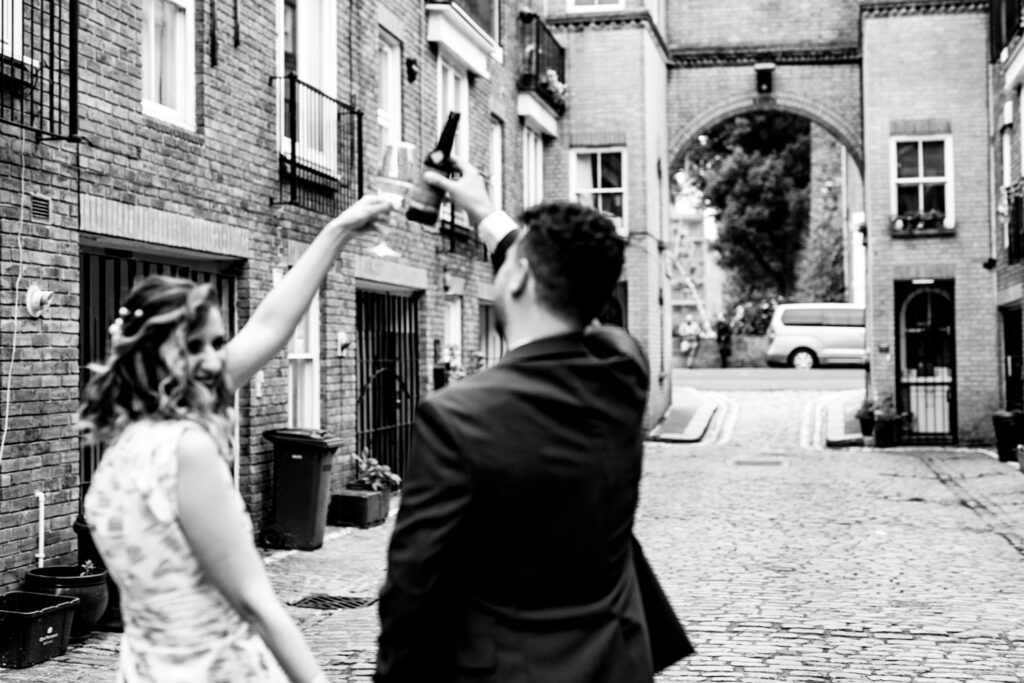 A bride and groom on their wedding day with a drink, walking together away from the camera.