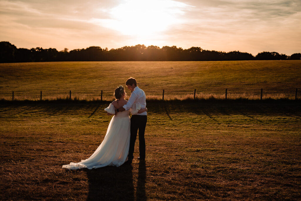 A couple stand in a field in front of a sunset in summer. The bride wears a flowing, white, festival style dress. 
