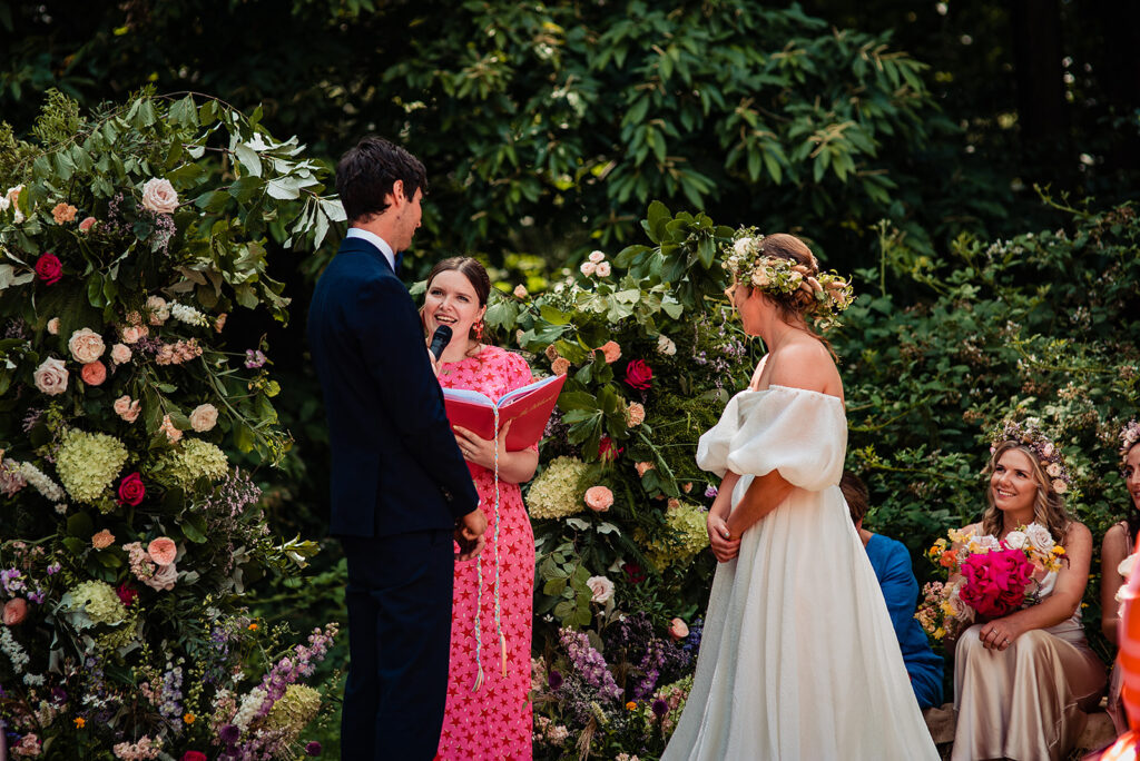 A summer festival wedding at Frickley Lake: a bride and groom say their vows with the celebrant, in front of statement installation florals
