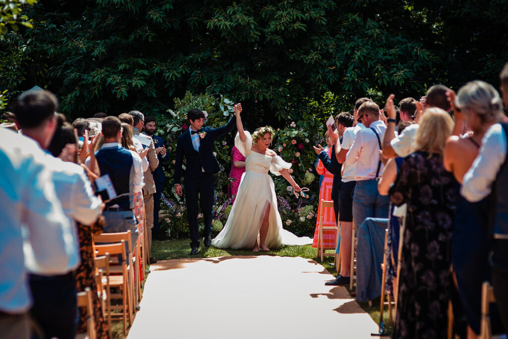 A summer festival wedding at Frickley Lake: A bride and groom joyfully walk back down the aisle after saying their vows. The guests stand and applaud. The wedding is an outdoor summer wedding. 