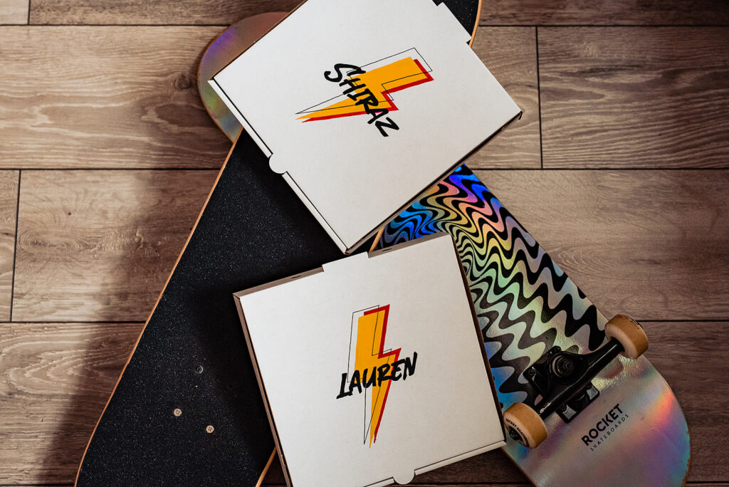 Two skateboards with alternative pizza boxes layed on top. The pizza boxes have Shiraz and Lauren written on them with a lightning bolt.