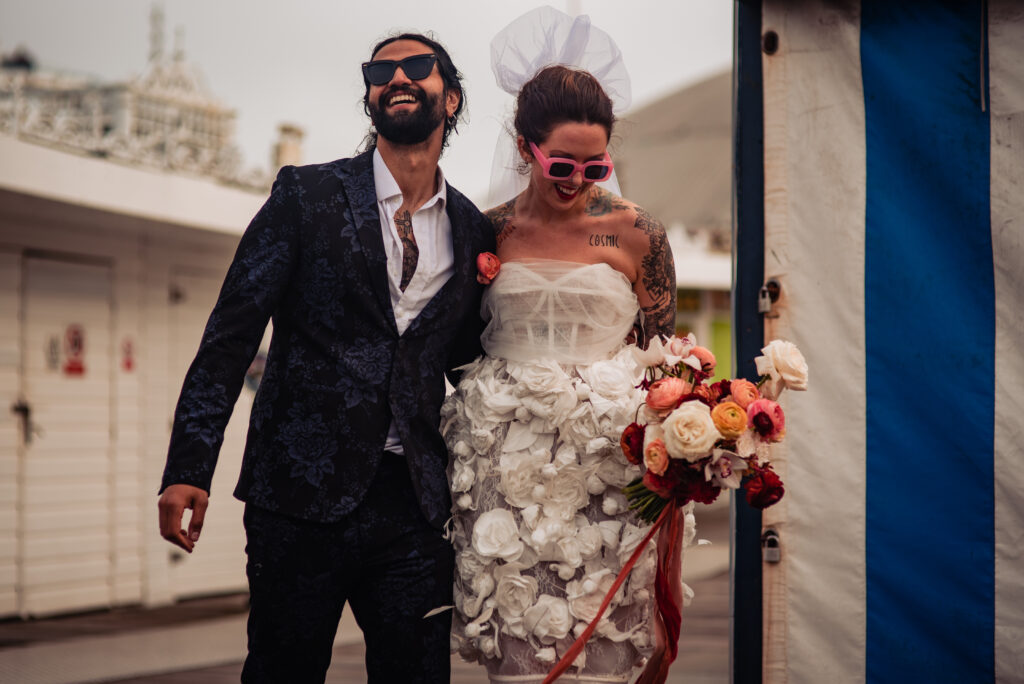A couple laugh on their elopement shoot. They both wear sunglasses and have an alternative look to their suit and dress. 