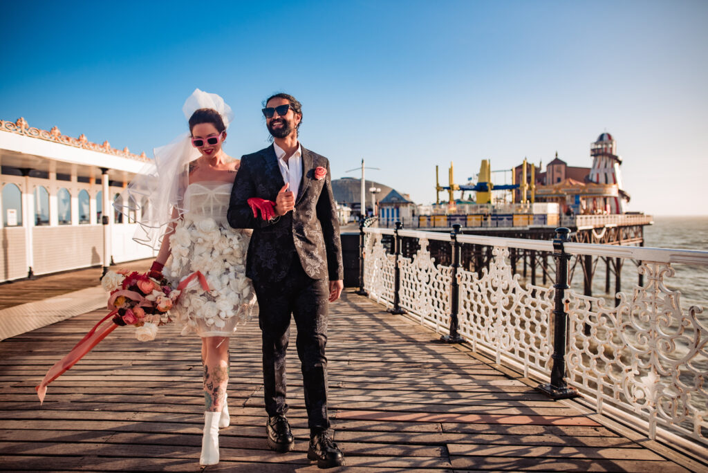 A bride and groom walk down the pier in a styled shoot in Brighton. The bride has tattoos and red leather gloves. and they both wear sunglasses. 