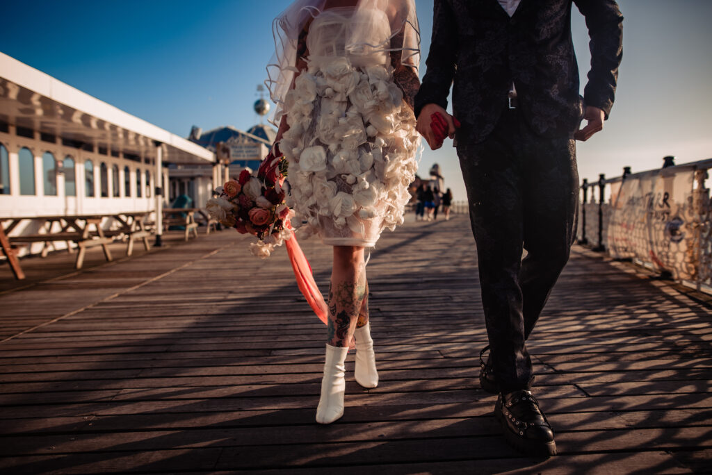 A couple walk down Brighton Pier holding hands. The bride has tattoos and white heeled boots.