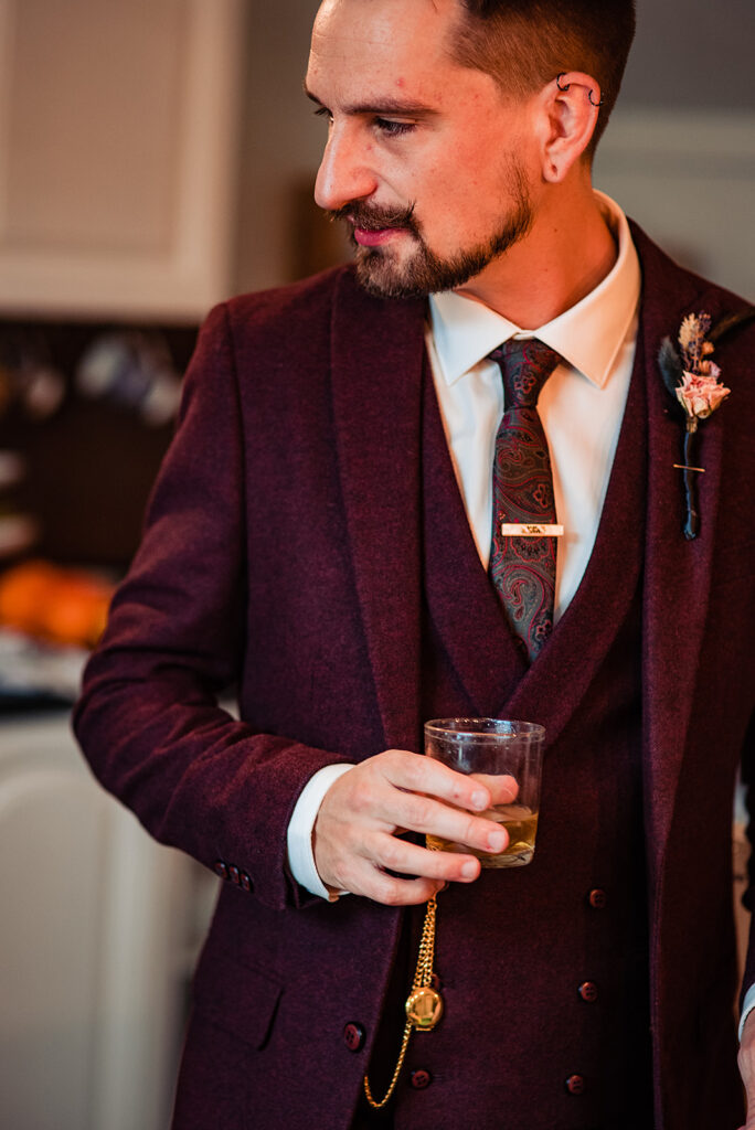 The groom wears a burgundy coloured suit with a waistcoat and buttonhole. He holds a drink and has a gold watch chain to accessorise his attire. 