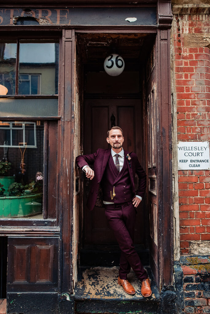 The groom leans in the doorway, looking outside. He wears a burgundy coloured suit with a waistcoat and buttonhole, he has a gold watch chain to accessorise his attire. 