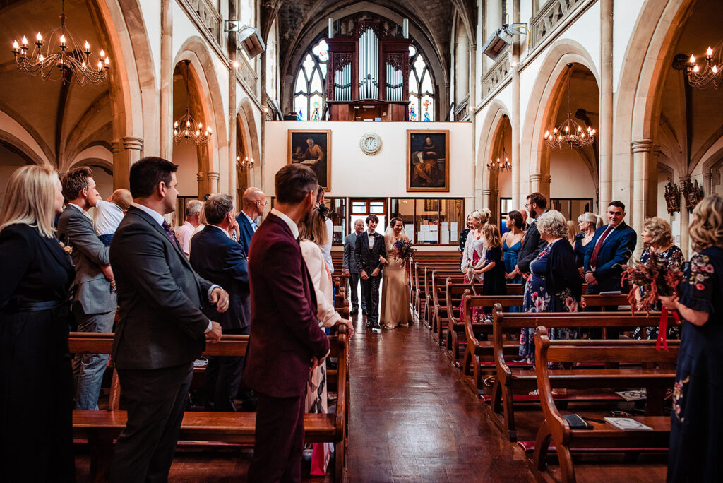 The bride walks in down the aisle of the church. She smiles at the guests who have turned to face her. The groom of this wedding is in the burgundy suit, looking back at his bride. 
