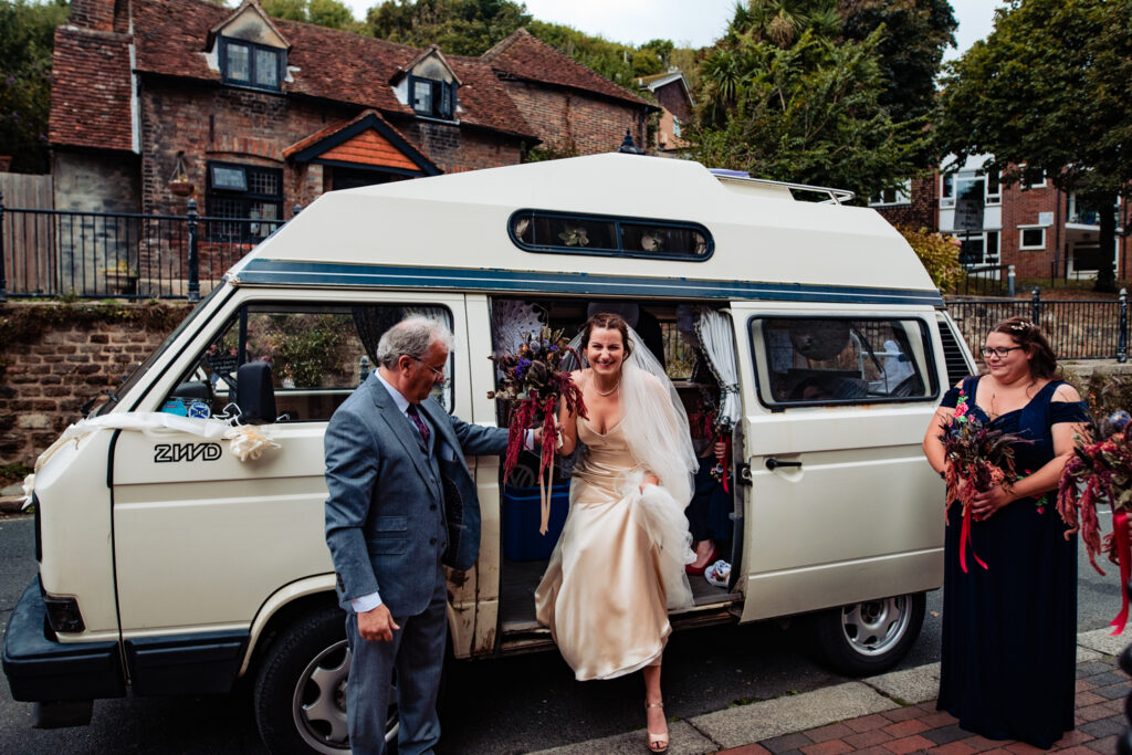 The bride climbs out of the wedding transport - an old cream camper van, decorated with ribbons. She smiles and lifts her skirt to climb out. 