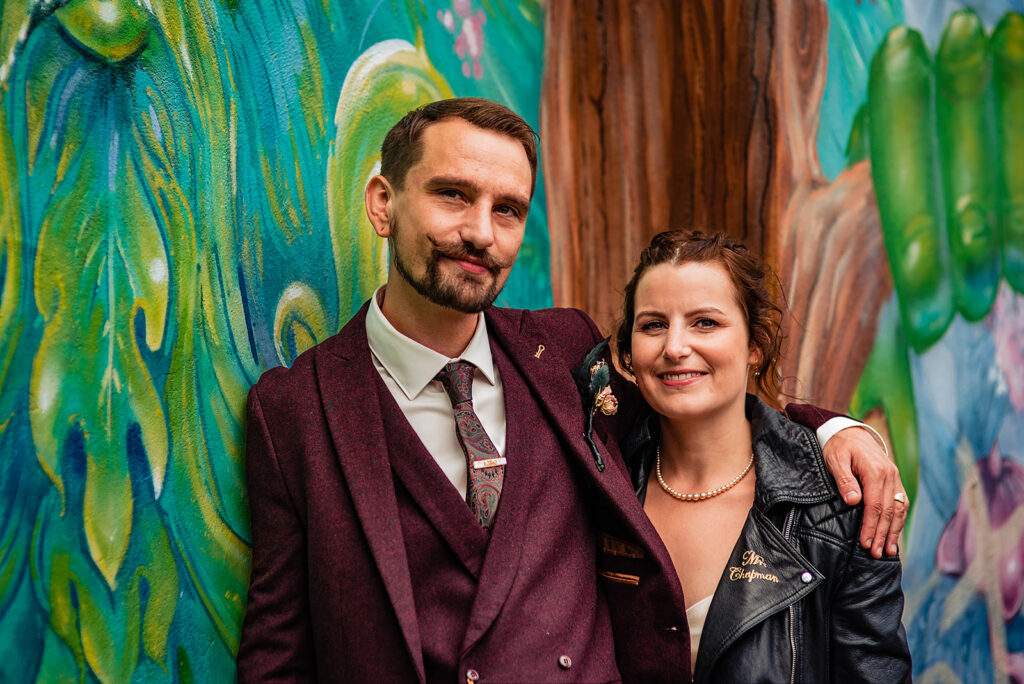 An Alternative wedding: Details from a couple's shoot with a bride and groom after their wedding ceremony. The bride wears a leather jacket in a goth chick vibe. The groom has his arm around her and they smile in front of a graffiti wall.