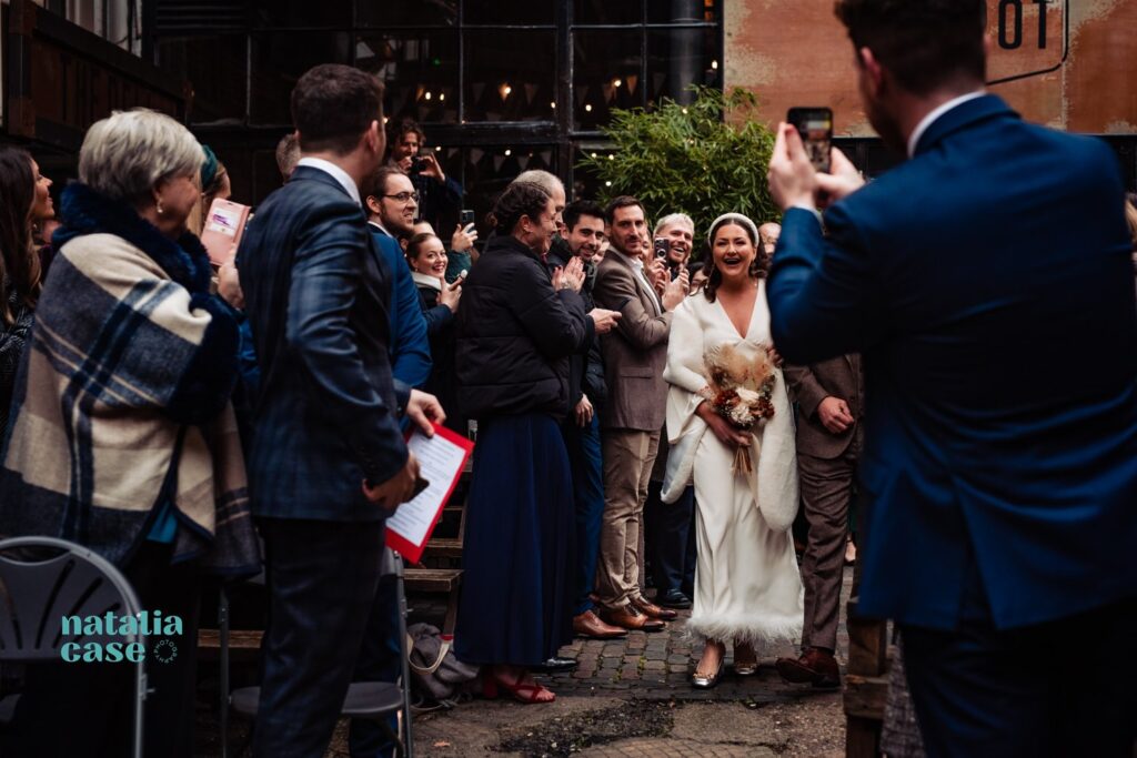 A bride and groom walk back down the aisle while guests take photos at The Depot N7, a London wedding venue. 