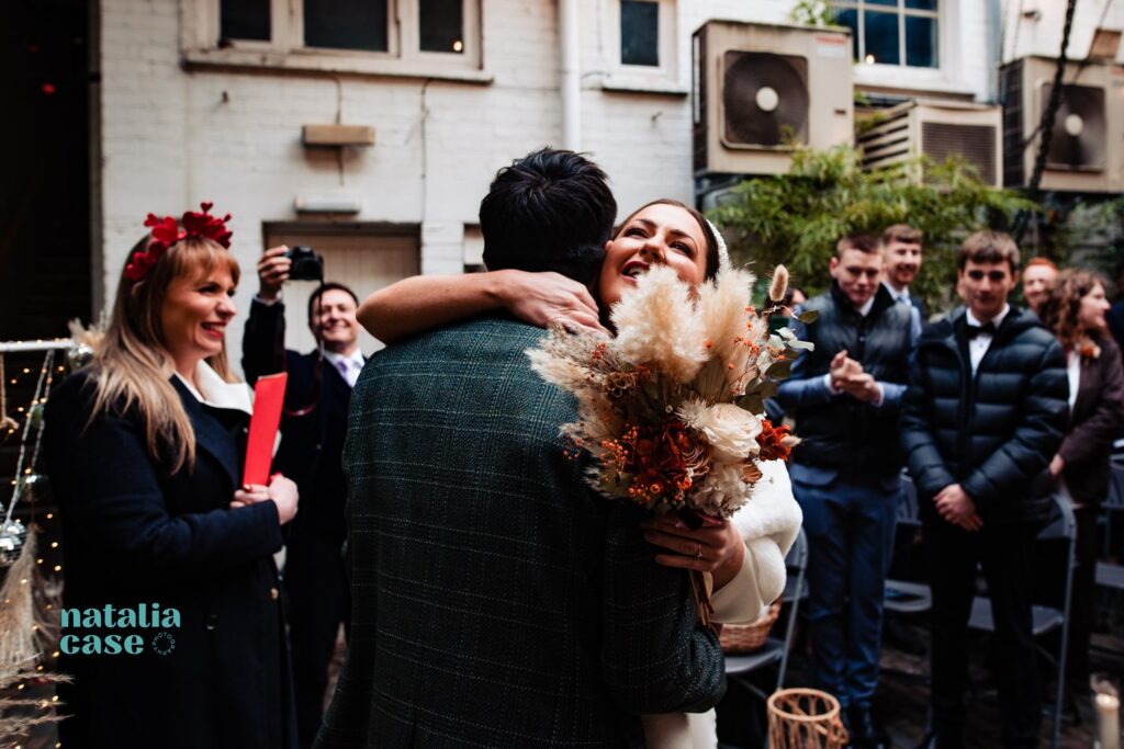 A bride and groom hug at the end of the ceremony, while the celebrant smiles and guests take photos. 