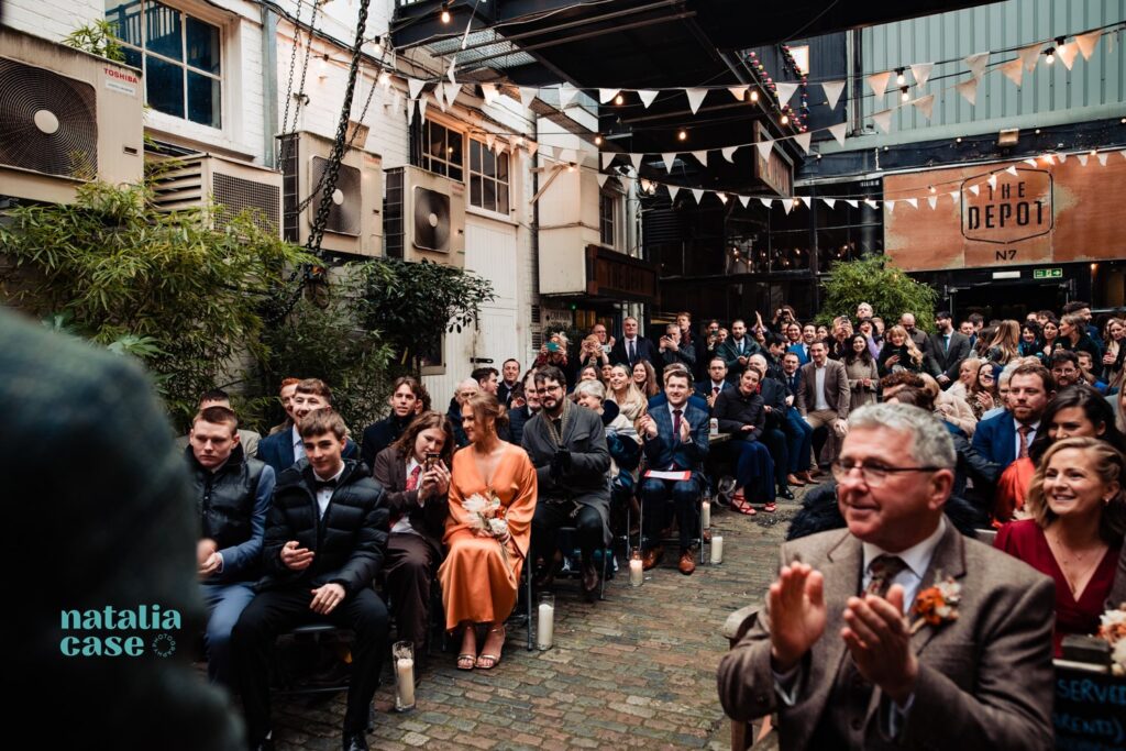 An outdoor wedding ceremony at The Depot N7 in London. Guests clap as they sit and wait for the ceremony to negin. 