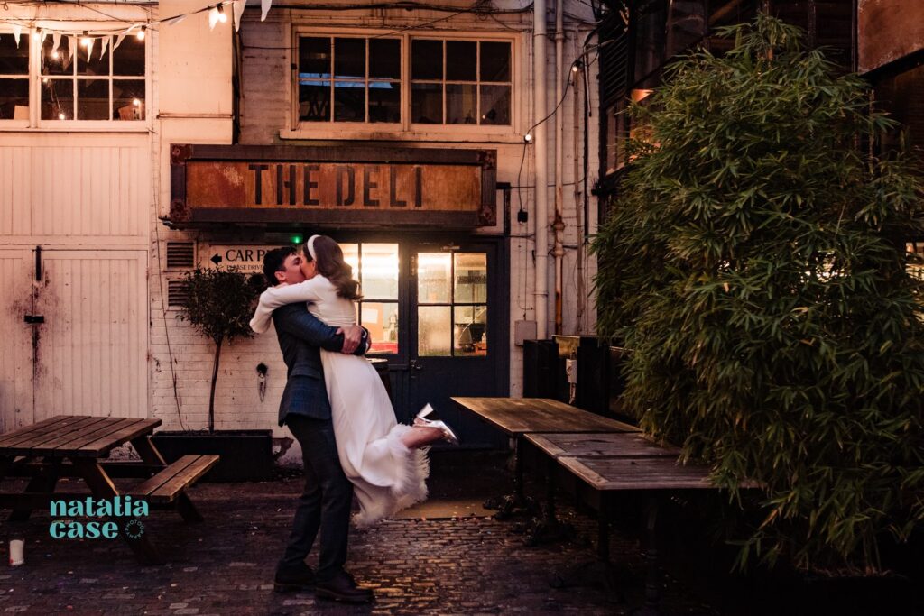 The bride and groom hug and spin in front of the 'Deli' sign at The Depot, N7. 