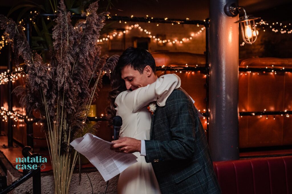 A bride and groom deliver their speeches to their guests at their winter wedding reception at the London venue, The Depot N7. The room is decorated with fairy lights. 