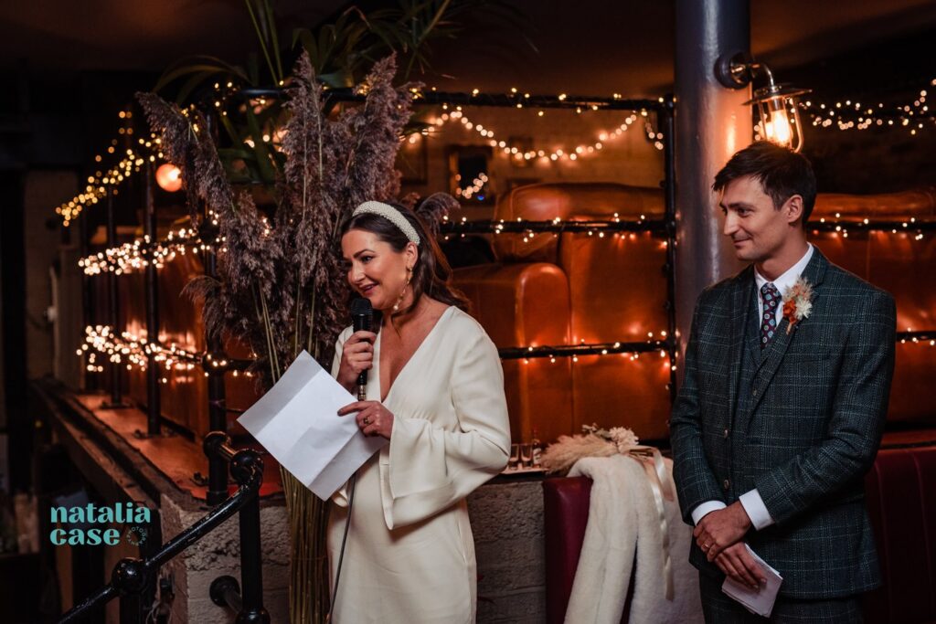 A bride and groom deliver their speeches to their guests at their winter wedding reception at the London venue, The Depot N7. The room is decorated with fairy lights. 