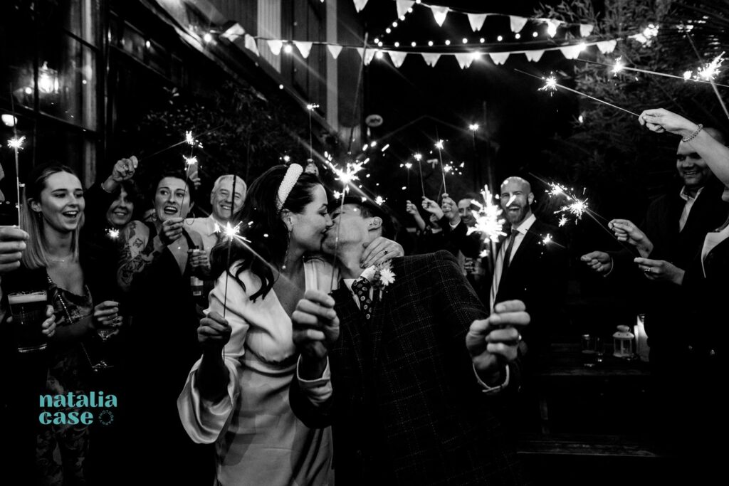 A bride and groom walk through and aisle of sparklers held by their guests, at the evening reception at their winter wedding. They kiss as they come closer to the camera.