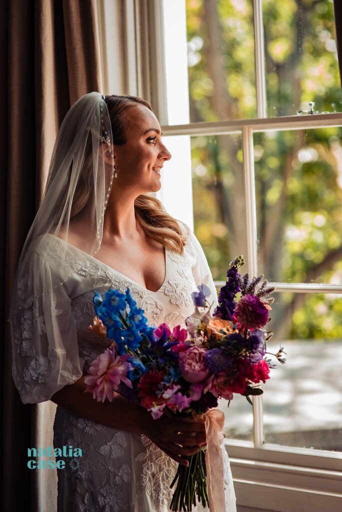 A bride looks out of the window after prepping for her wedding. She is staying in the Bingham RIverhouse, Richmond, where the wedding will take place. She holds a colourful bouquet. 