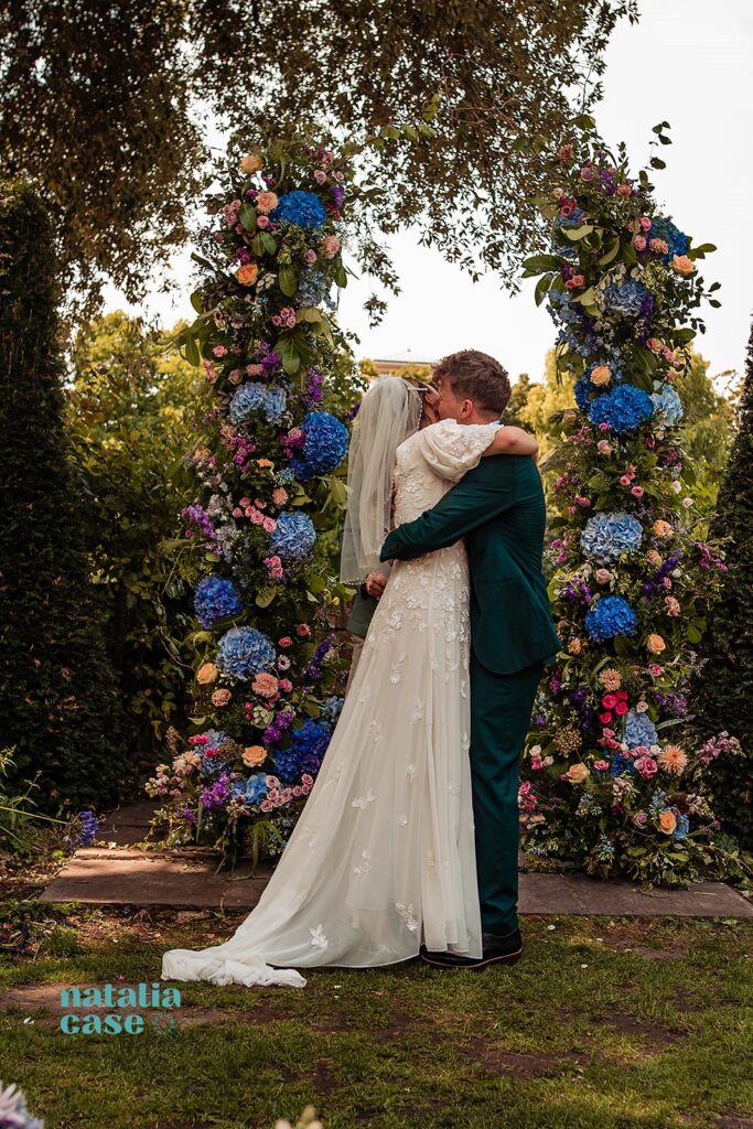 A bride and groom embrace under a flower arch at their outdoor summer wedding