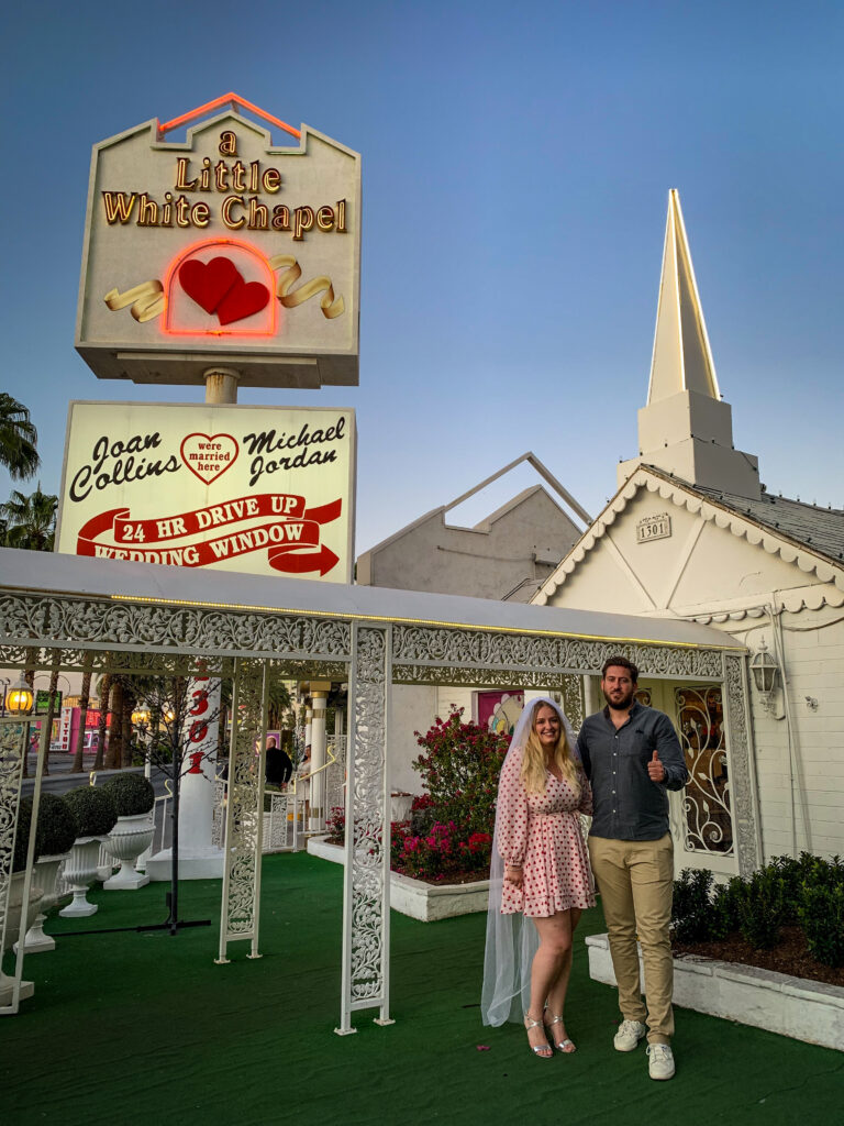 A bride and groom stand outside an elopement wedding venue in Vegas, USA. She wears a short pink polka dot dress and long white veil, and he shows a thumbs up to the camera. 