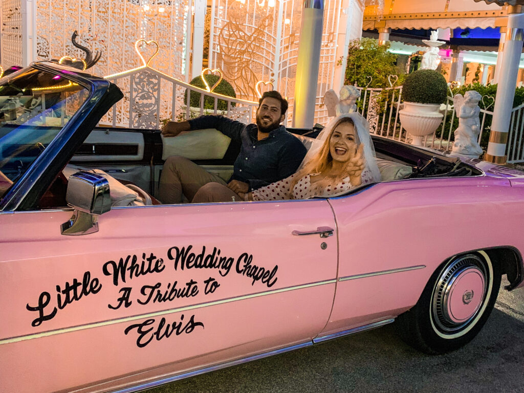 A bride and groom smile for the camera. They sit in a pink classic American car after their Vegas elopement wedding ceremony. 