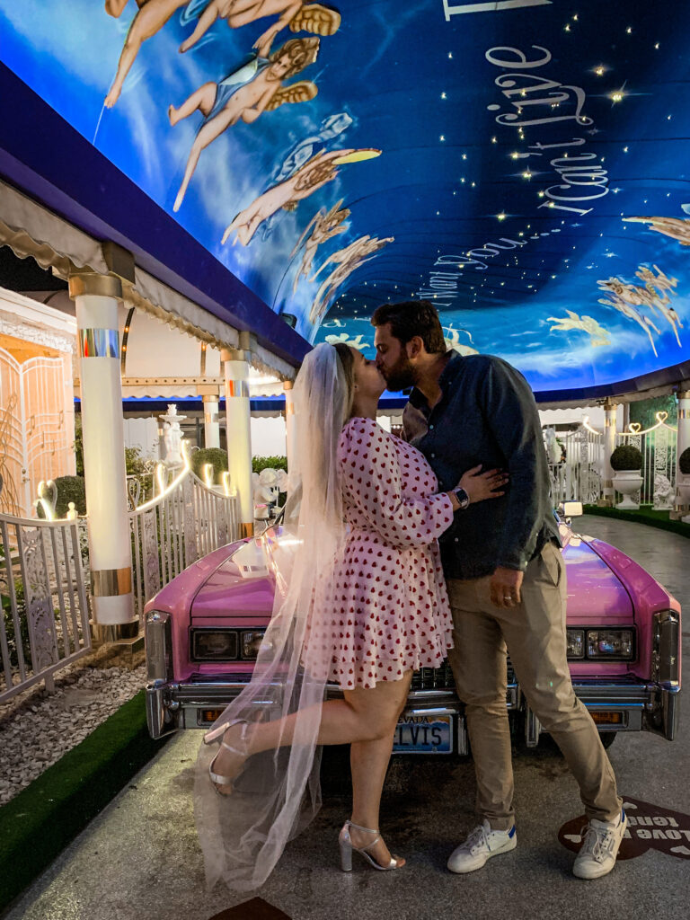 A bride and groom kiss outside an elopement wedding venue in Vegas, USA. She wears a short pink polka dot dress and long white veil, and he wears a dark shirt. The ceiling is decorated in a dark blue design with angels painted on. 