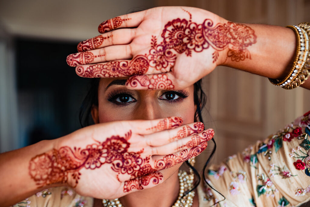 The bride's bridal henna on her fingers and palms. She's holding her hands up to cover her face, except her stunning eyes.