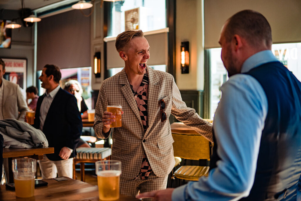 A groomsman laughing with another guest, holding a pint in a London pub