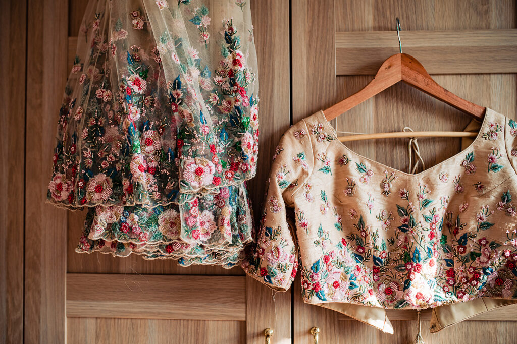 Traditional Hindu wedding attire, hanging up against a light wood wardrobe. The top and skirt are cream but covered in colourful embroidered flowers of pink, red, turquoise, green, and blues. 