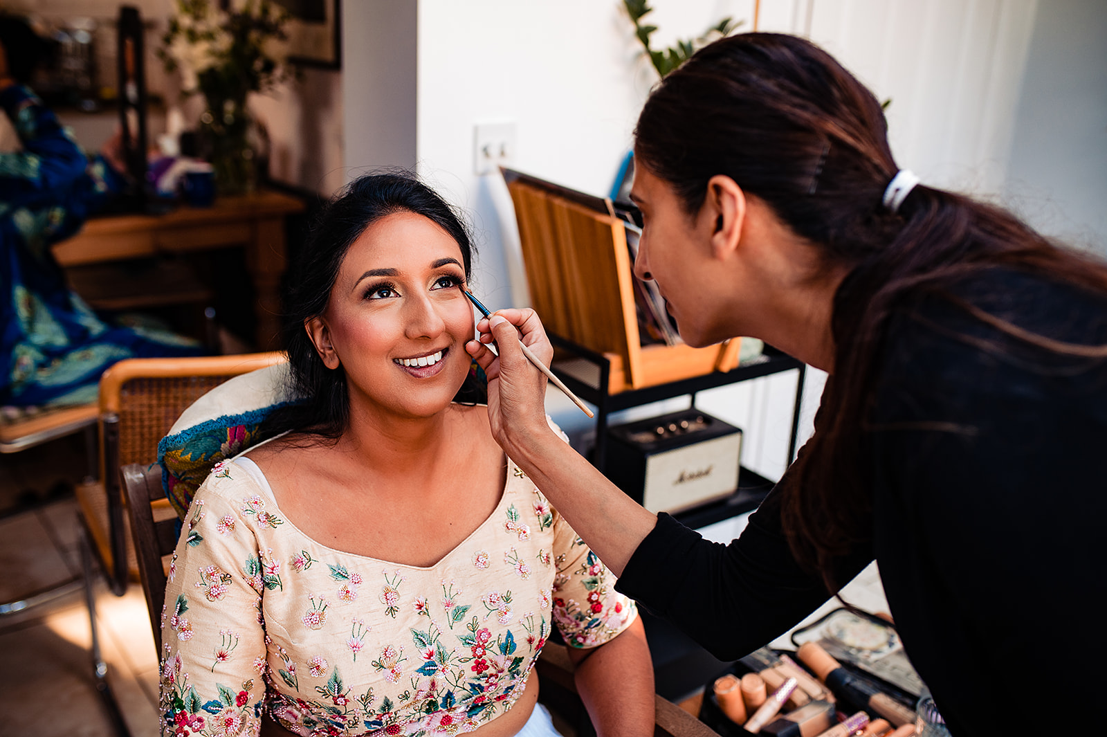 The bride gets her makeup done, and is beaming. She is sat down with her traditional Hindu outfit on that is adorned with embroidered flowers of all different colours. The lighting is bright and summery.