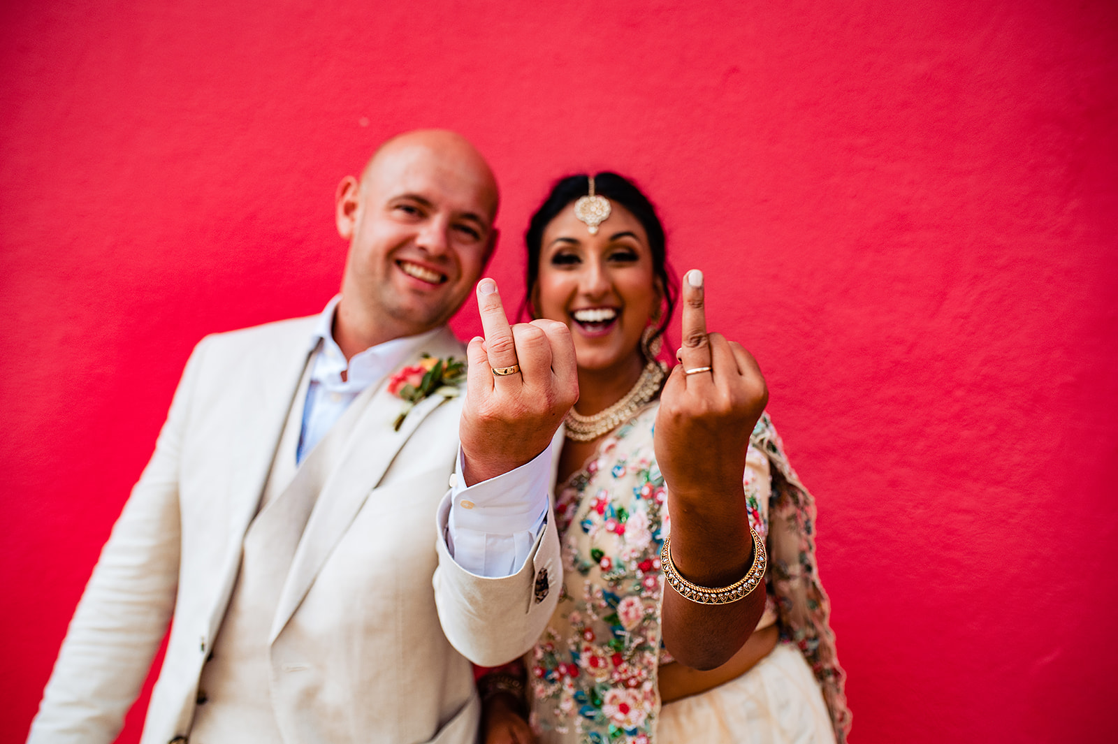 The bride and groom stand against a super bright crimson wall, holding up their ring fingers as if they're flipping the bird. Ollie, the groom, is wearing a cream linen suit with no tie, and a bright orange contrasted button hole flower. Krishna, the bride, is adorned in traditional Hindu attire with bright, colourful flowers embroidered on. They're both smiling from ear to ear.