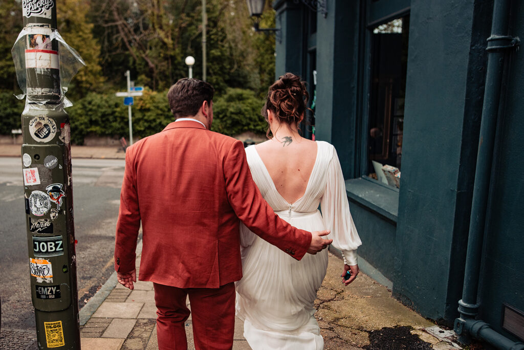 A married couple facing away from the camera. The groom is wearing a burnt orange suit, and the bride is wearing a white dress.