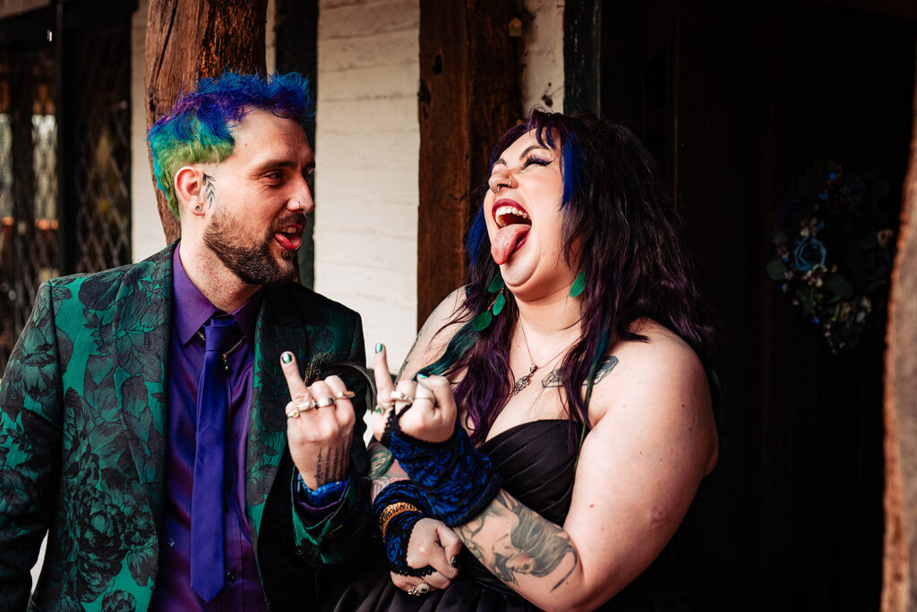 An alternative couple posing for the camera. The bride and groom show off their wedding rings and wear green and purple wedding attire.