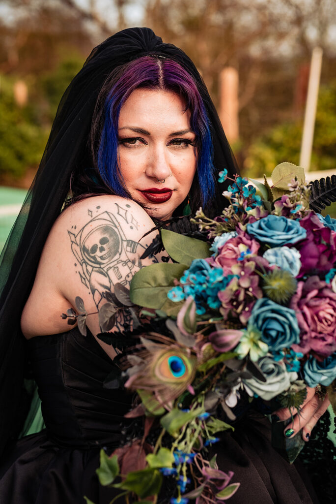 a close-up portrait of an alternative bride. She is wearing a black dress, with a black veil. In her hands, she is holding a bouquet.