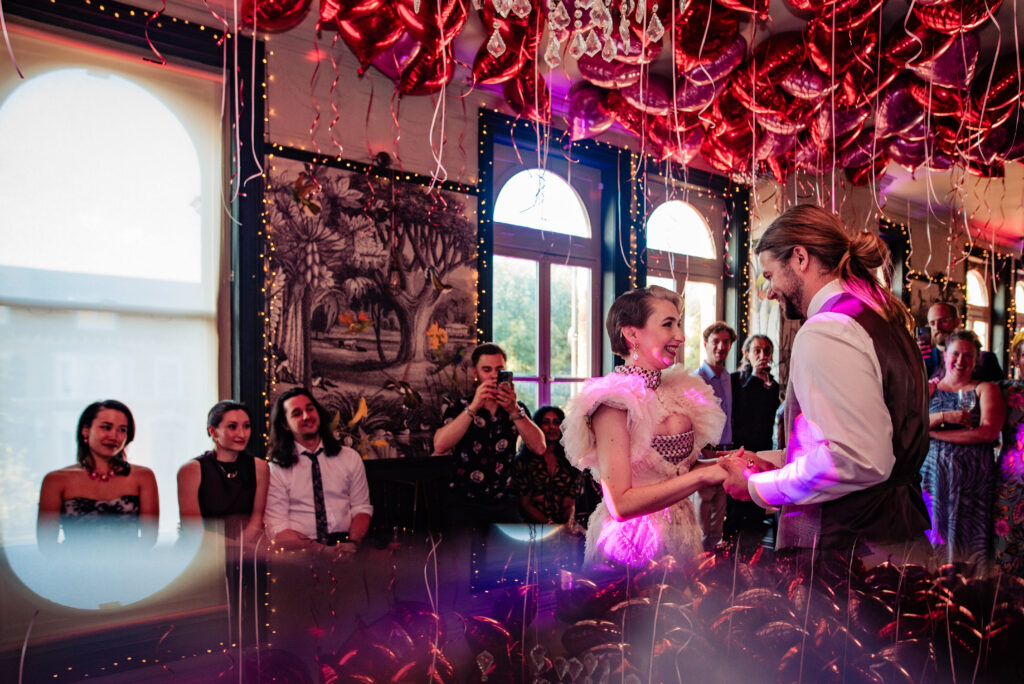 A wedding couple, surrounded by people watching them do their first dance. There are pink and red heart shaped balloons on the ceiling. The couple are holding hand and dancing.