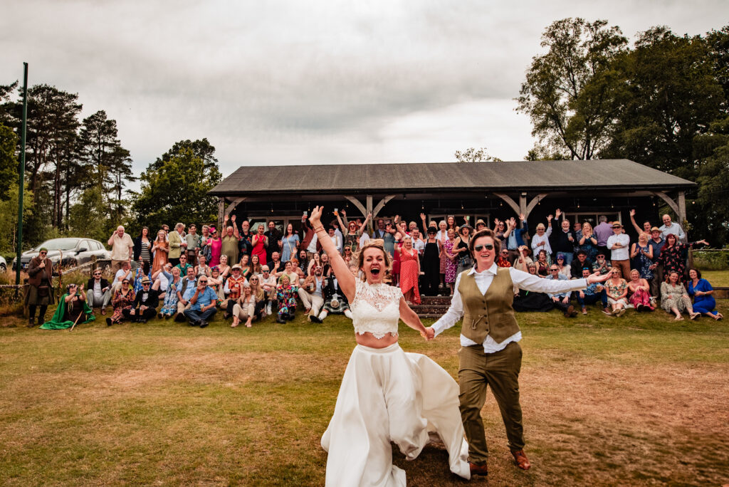 A group photo of a wedding guest sitting in front of a Cricket Club. Two brides, one wearing a white dress and one wearing a suit are running towards the camera.